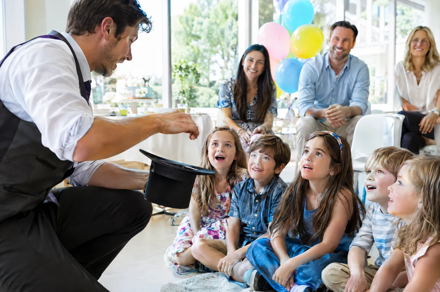 Kids’ party: a magician shows his hat to children sitting on the floor, parents behind them