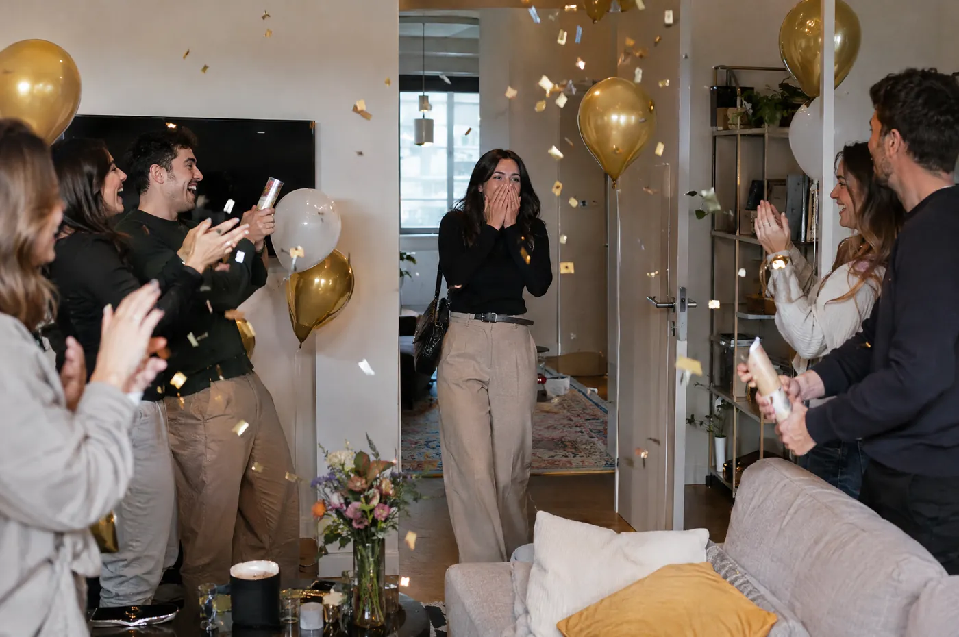 Surprise party: she walks into the living room, hands over her mouth, under confetti and gold balloons