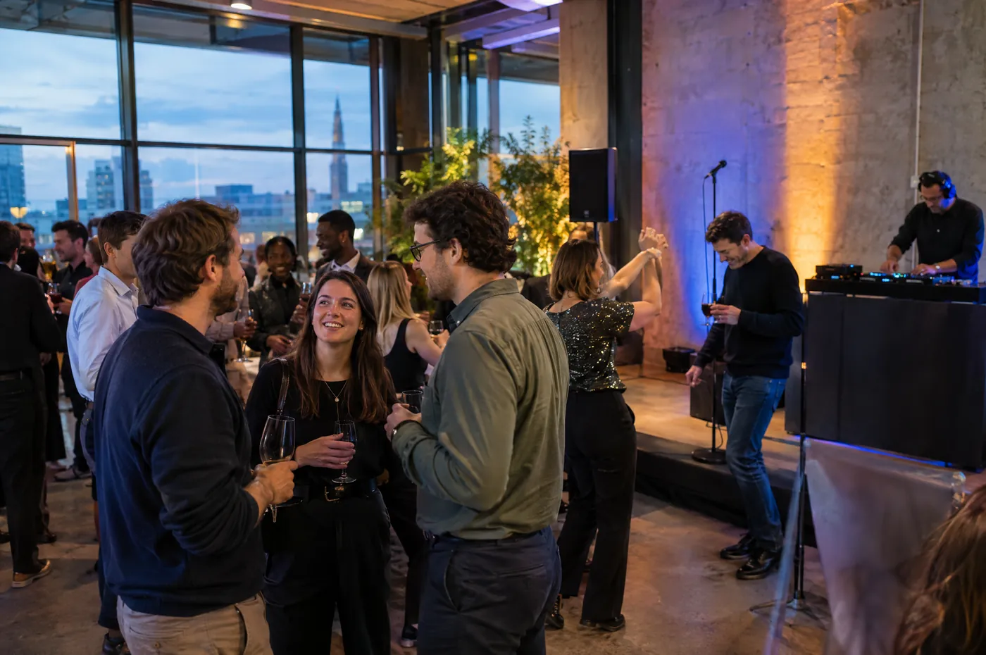 Soirée d’entreprise dans un espace en béton brut avec vue sur la ville, un DJ joue sur une petite scène