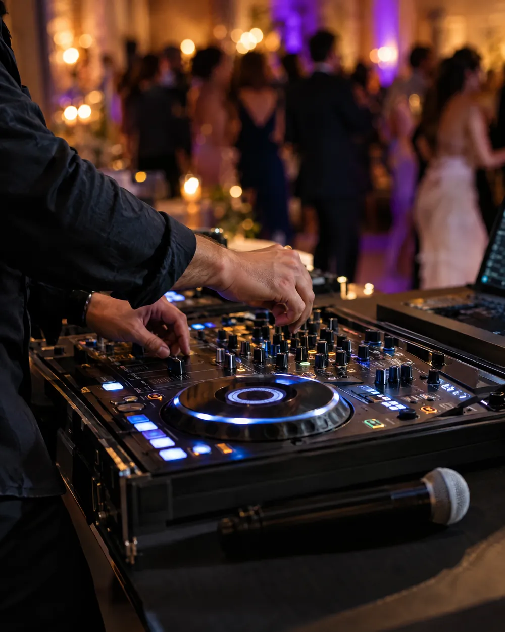 Les mains d’un DJ sur sa table de mixage, un micro sans fil posé devant, la piste d’un mariage floue au fond